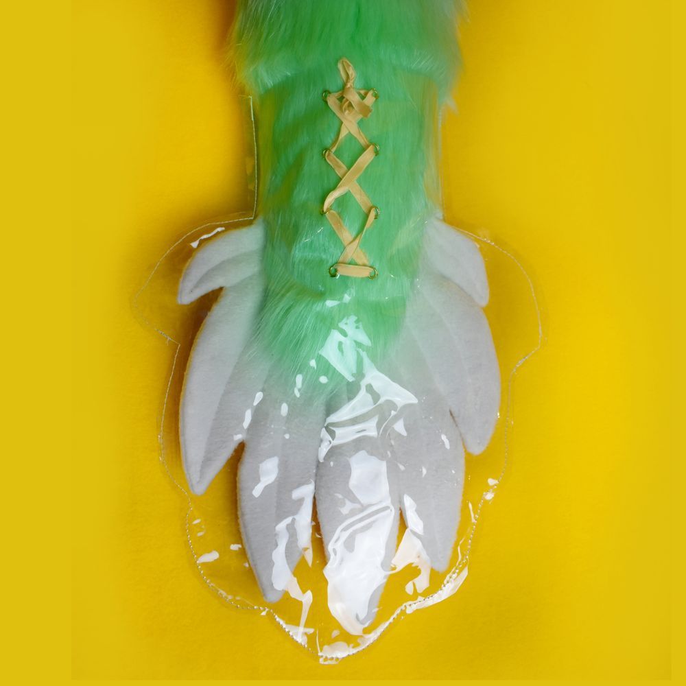 Close-up photo showing details from a set of commissioned items made for a furry buyer by A Kindness of Crows. The tip of a giant dragon tail is visible - made from mint green luxury faux fur, with a white fleece feathered tip. The tip of the tail is protected by a clear plastic cover, laced up with a caramel satin ribbon. The background is vivid yellow.