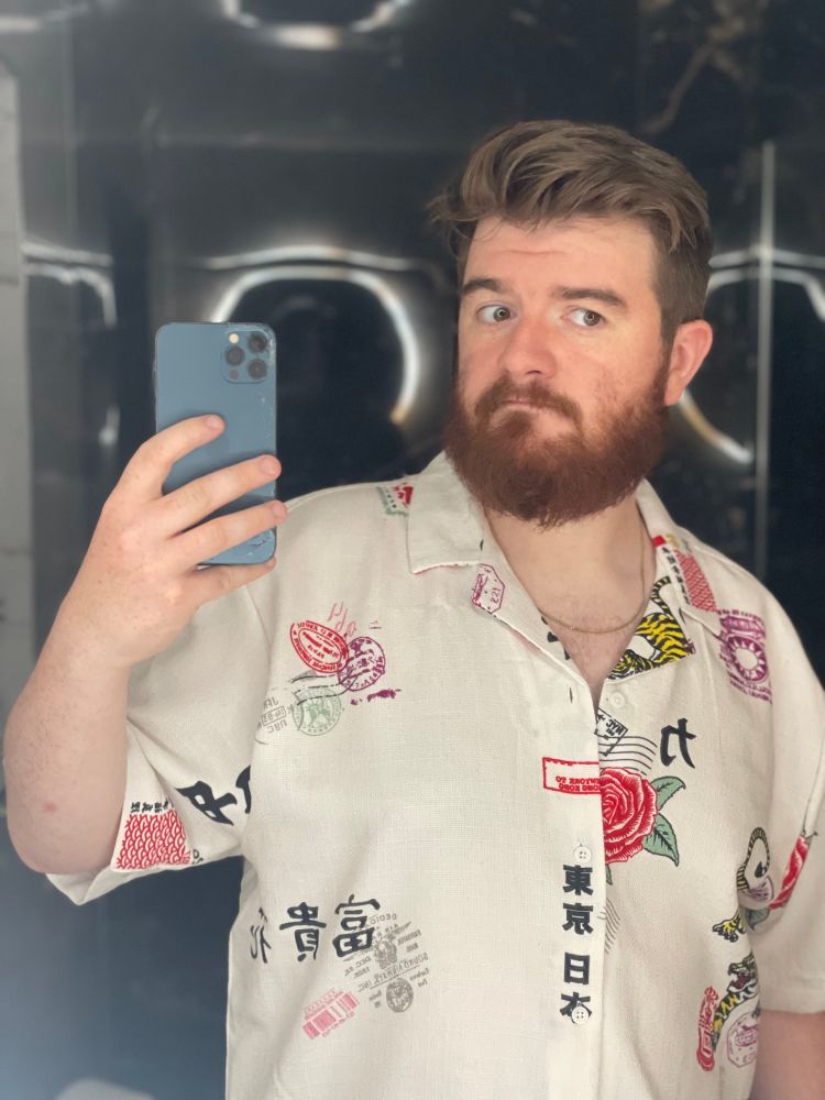 Photo of a man with beard taking a selfie, wearing white linen-esque shirt with some tiger and postmark symbols 
