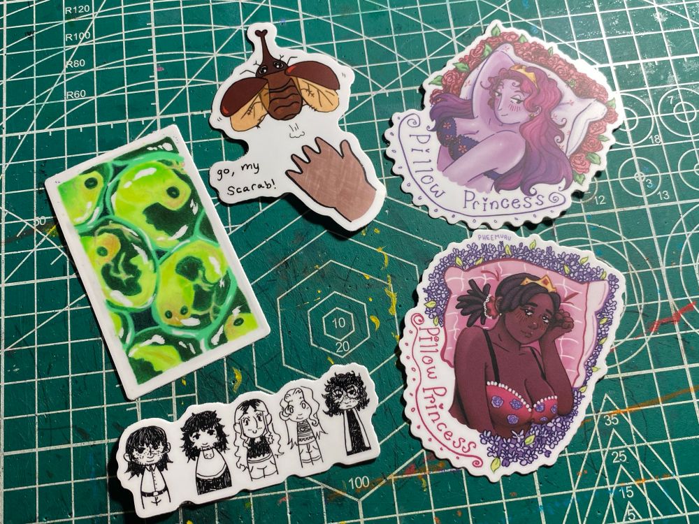 5 stickers
- 2 pillow princess stickers, feminine figures in bras laid back on a pillow. One sticker is a non black woman and the other is a black woman
- HNTF cast chibi sticker 
- Tadpole egg sticker
- "go, my scarab!" sticker with a beetle taking flight from a hand