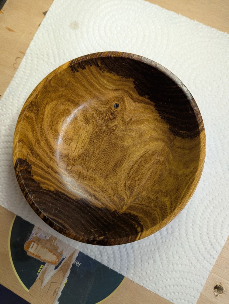 A photograph of a hand turned laburnum bowl sitting on a paper towel, on a lathe bed. The bowl is two tone, with two dark ovals opposite each other, and the rest a lighter colour with striking grain lines. It's hard to see in this image, but there's torn grain throughout the dark sections.