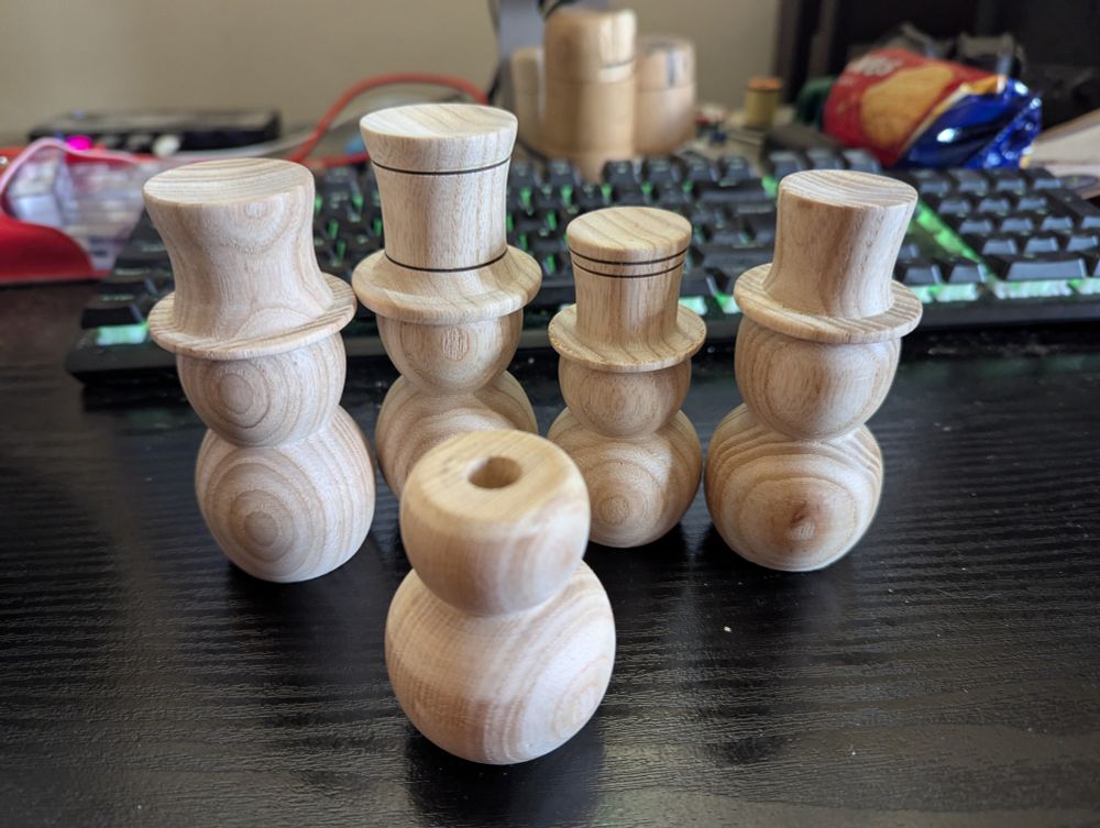 A photograph of a black desk on which sit four complete snowmen and a fifth without a hat. The fifth shows how I drill a 10mm diameter hole in the head in the head to locate a matching pin on the bottom of the hat, which is turned separately.