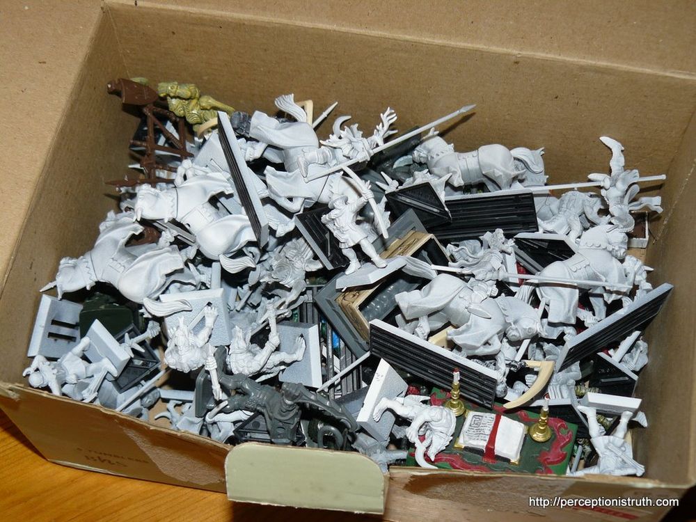 A photograph of a cardboard box, filled with plastic fantasy miniatures. A mixture of knights on horseback, knights with bows, lizardmen, and some random pieces of furniture from a hero quest board game.