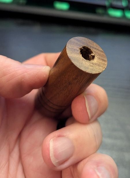 Photograph showing a hand, holding the bottom of a hand-turned wooden needle case. The base has a hole all in it, and it should not.