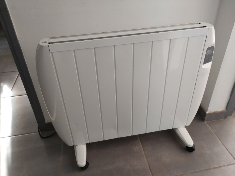 Un radiateur électrique blanc à inertie sèche posé sur ses roulettes dans un couloir entre deux passages de portes, on y voit un panneau de commande éteint sur son côté droit. Il semble attendre qu'on lui donne un prénom.