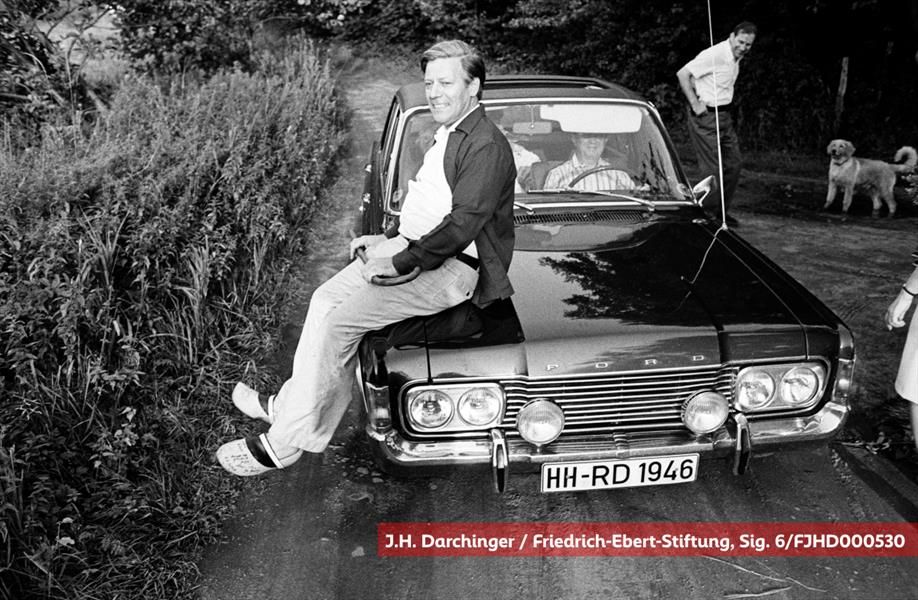 Helmut Schmidt sitzt grinsend auf der Motorhaube eines Ford, am Steuer ein Mann, im Hintergrund ein weiterer, lachend