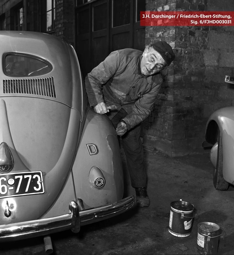 Schwarz-Weiß-Foto eines Arbeiters in schmutziger Arbeitskleidung, der eine Beule aus einem VW Käfer hämmert