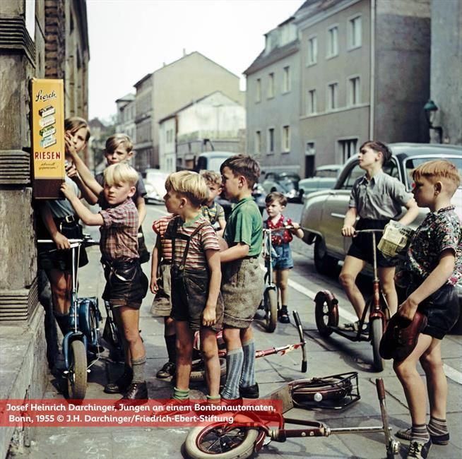Szene von 1955, eine Gruppe von Jungen mit Tretrollern steht vor einem Automaten mit der Aufschrift "Storck Riesen" 