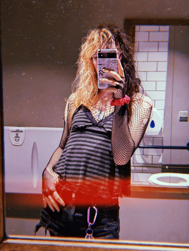 mirror selfie of myself in a disabled toilet wearing an outfit consisting of fishnet sleeves, stripped camisole top and black shorts with a large purple carabiner :3
