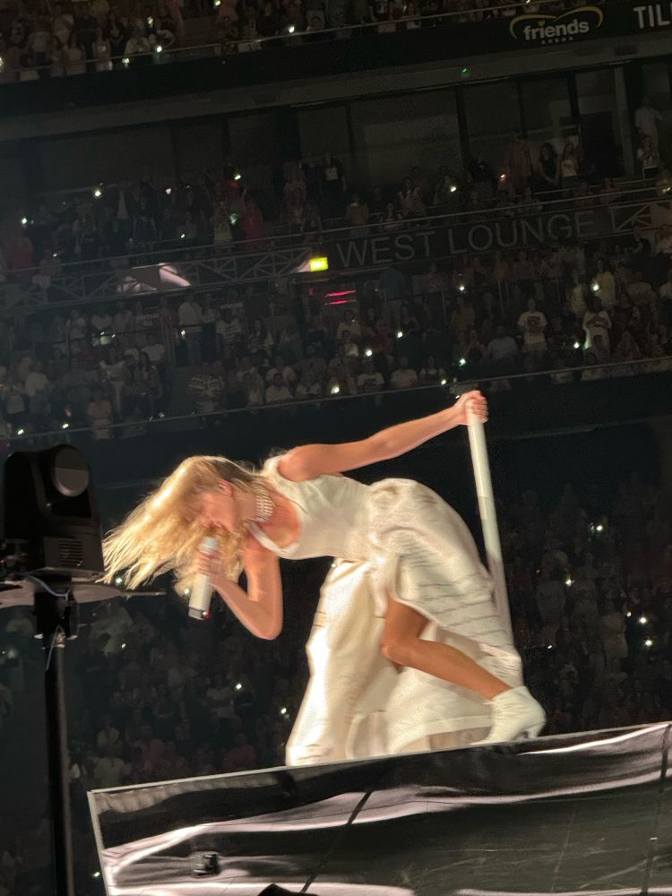 "Taylor Swift singing on stage at the Eras Tour, wearing a sparkly outfit and surrounded by cheering fans during the TTPD era." 