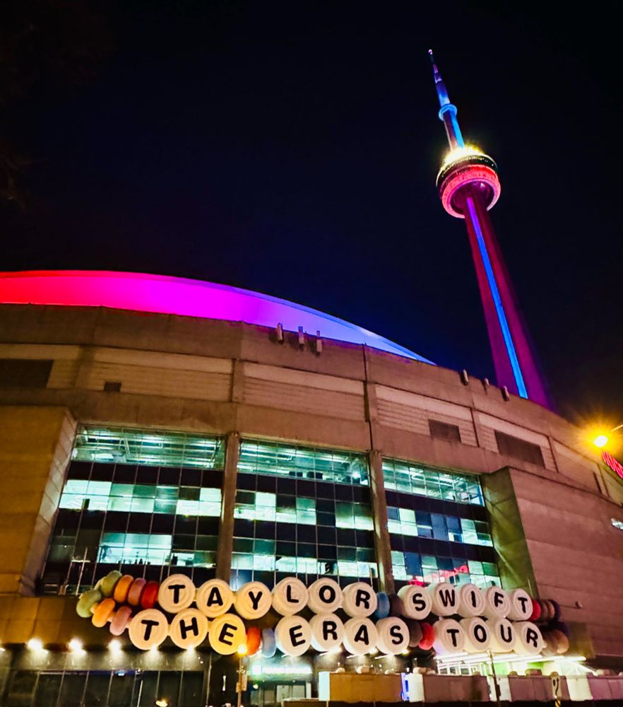 A night shot of the Rogers Centre in Toronto, illuminated in pink and blue lights. The CN Tower, lit up in blue and white, stands tall in the background. A large banner with the words "TAYLOR SWIFT THE ERAS TOUR" hangs across the front of the stadium.