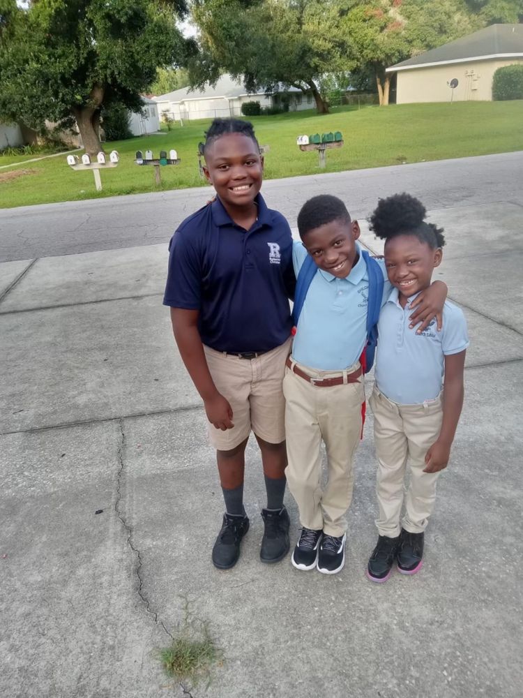 Isaac, Israel e Afrika Owens, todos de calça caqui e camisa polo azul de uniforme, sorriem para a câmera. Isaac é o maior, cheinho e com o cabelo trançado pra trás, Izzy tem fade nas laterais e cabelo curtinho em cima e Afrika tem o cabelo em dois puffs, um no topo e um na parte de trás da cabeça. Os 3 sorriem largamente, abraçados 