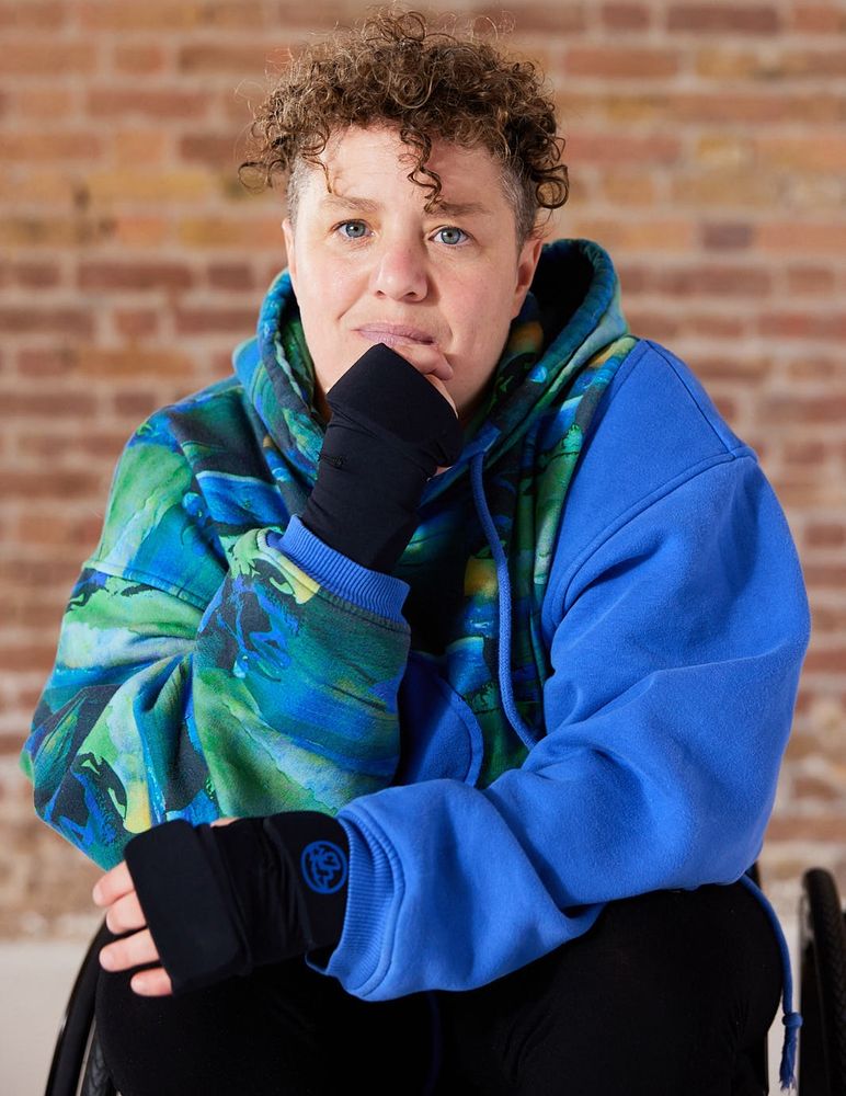 A photo of Jess Thom, a white, wheelchair using woman looking into the camera with a serious expression. She is wearing a blue and green hoodie, black padded gloves and is pictured in from of a brick wall