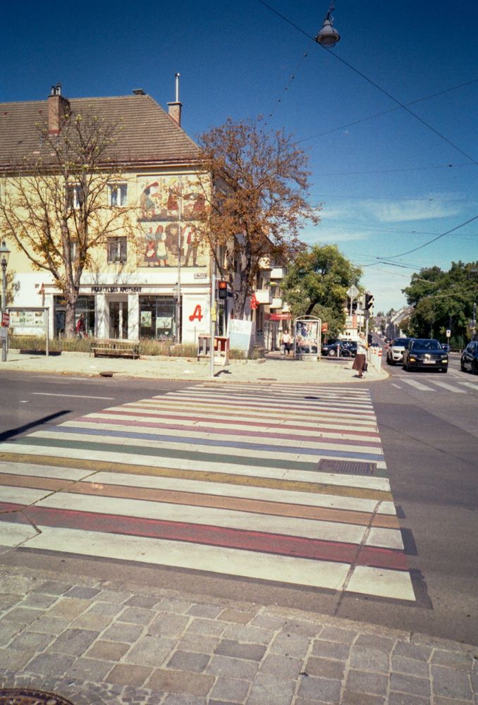 Auf dem Foto ist eine Straßenkreuzung in einer Stadt zu sehen. Im Vordergrund befindet sich ein Zebrastreifen, der nicht wie üblich schwarz-weiß, sondern in verschiedenen Farben bemalt ist – die Streifen sind in Pastellfarben wie Rosa, Gelb, Grün, Blau und Lila gehalten.

Im Hintergrund sieht man ein gelbliches Gebäude mit einem Wandgemälde, das mehrere Figuren darstellt. Im Erdgeschoss befindet sich eine Apotheke, erkennbar am roten Apothekenzeichen („A“). Vor dem Gebäude stehen Bäume, die teilweise bereits herbstlich verfärbte Blätter tragen.

Rechts im Bild fahren mehrere Autos auf der Straße, und eine Person überquert weiter hinten die Fahrbahn. Über der Szene verlaufen Straßenbahn-Oberleitungen, und eine Straßenlampe hängt von den Kabeln herab. Links am Gehsteig steht eine Bank, daneben sind Werbeplakate sichtbar.
Die Szene wirkt sonnig, mit blauem Himmel und klarer Sicht.

(Text KI-generiert)