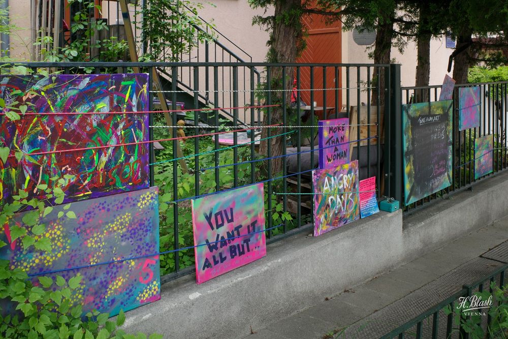 Das Bild zeigt einen Zaun vor einem Wohnhaus, an dem mehrere bunte, handgemachte Schilder und Leinwände aufgehängt sind. Die Werke wirken spontan und künstlerisch, teils im Stil von Graffiti oder Street Art.

Zu sehen sind u.a.:

    Links oben eine bunte, abstrakte Leinwand mit chaotischen Pinselstrichen, darauf der rote Schriftzug „insel“.

    Unten eine violette Leinwand mit bunten Punkten und der Zahl „5“ in Pink.

    Mittig ein Schild mit dem Text: „YOU WANT IT ALL BUT…“ in schwarzer Schrift auf rosa-grünem Hintergrund.

    Rechts oben ein pinkes Schild mit „MORE THAN A WOMAN“ in schwarzer Schrift.

    Darunter eine Leinwand mit „ANGRY DAYS“.

    Ganz rechts eine große Tafel mit der Aufschrift „TAKE AWAY ART“ und einer Liste von „PRICES: 0-10-20-30 EUR“ sowie der Anmerkung „Pay what you want“.

    Drumherum viel Grün, Efeu und Sträucher, sowie eine offene Treppe im Hintergrund.

Das Gesamtbild wirkt wie eine kleine Straßenkunstausstellung, bei der Passanten Kunstwerke mitnehmen können.