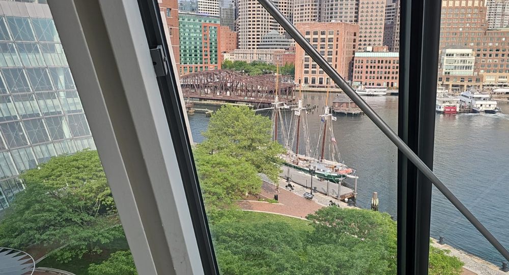 A three-masted schooner with a progress pride flag is tied up to the first circuit's dock in Boston. The photo is taken from inside the building looking out at the building's private garden and the river. Boston's downtown is in the background.
