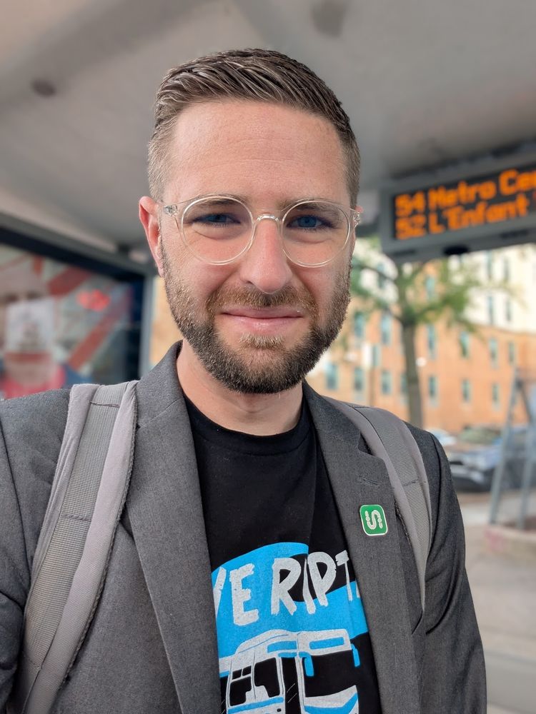 Photo of me at a bus stop in DC wearing a black "Save RIPTA" shirt beneath a grey blazer with a Transit app pin