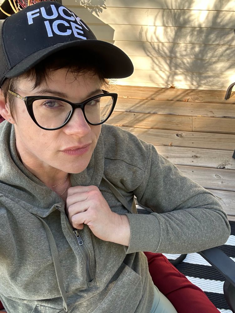 Women with black glasses and light green hoodie wears a black baseball cap with the words FUCK ICE on it.