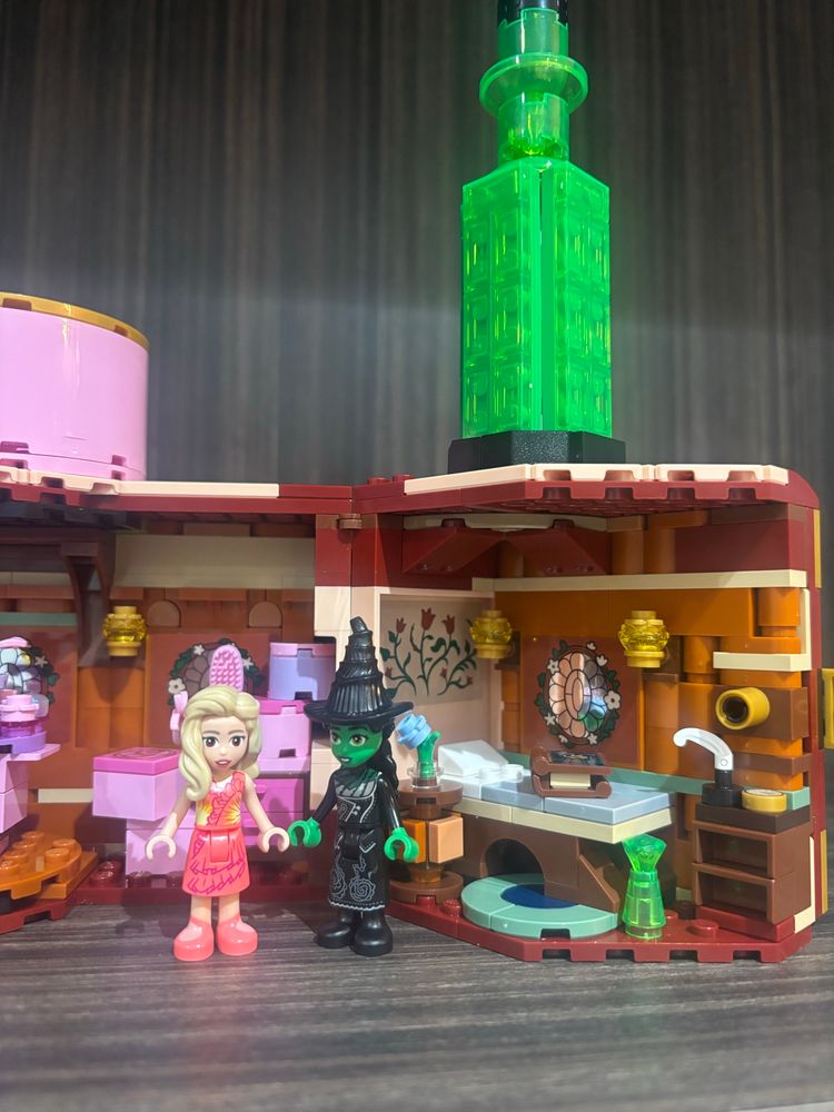 the inside of a lego doll house based on the wicked movie. this picture shows elphaba’s side of the dorm room and shows the glinda mini doll in an orange and yellow outfit with a pink flower in her hair and the elphaba mini doll wearing a black outfit and wearing a witch hat
