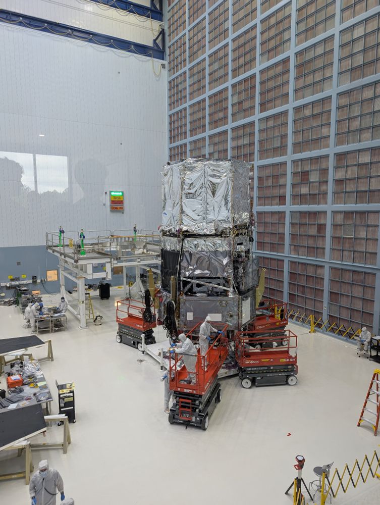 The Roman space telescopes main instrument package and superstructure is surrounded by engineers in bunny suites in a giant clean room. Behind the telescope is a huge wall of filters cleaning the air.