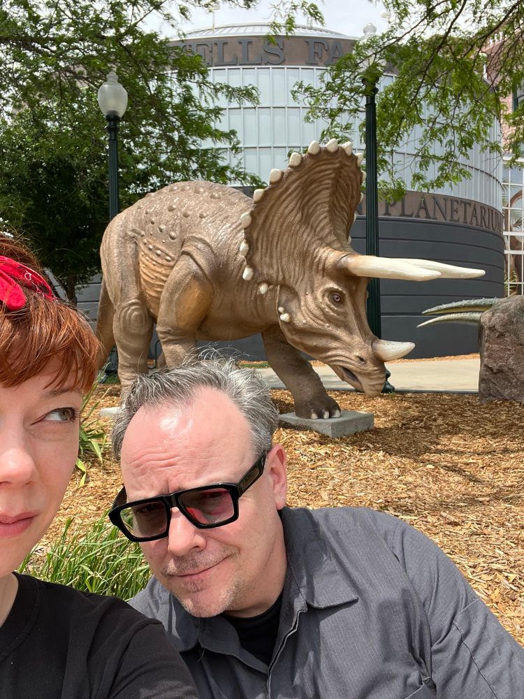 A doofus takes a selfie that includes half of her face on the left side of the frame, her handsome husband squinting in the sun as his transition lenses darken, and a triceratops sculpture in the background at the Washington Pavilion.