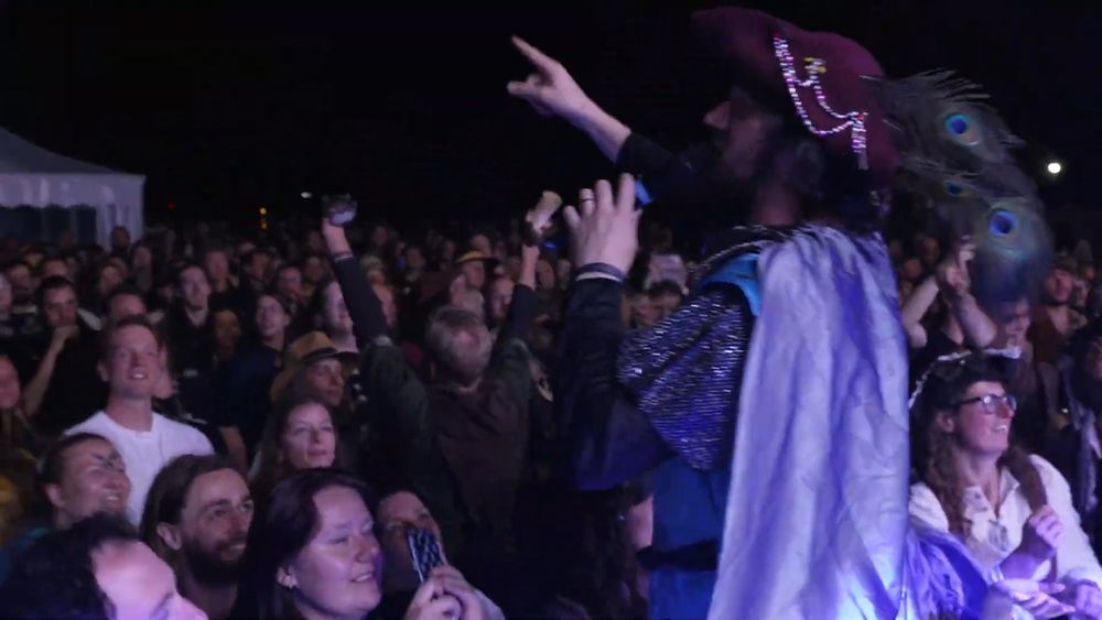 Longest Johns singer Robbie stands on the barrier at CastleFest fantasy festival,  in the middle of their set. He's wearing a knight's tabard and short cloak and he has just stolen a fancy tricorn hat, with beads and a peacock feather, from a member of the audience.
