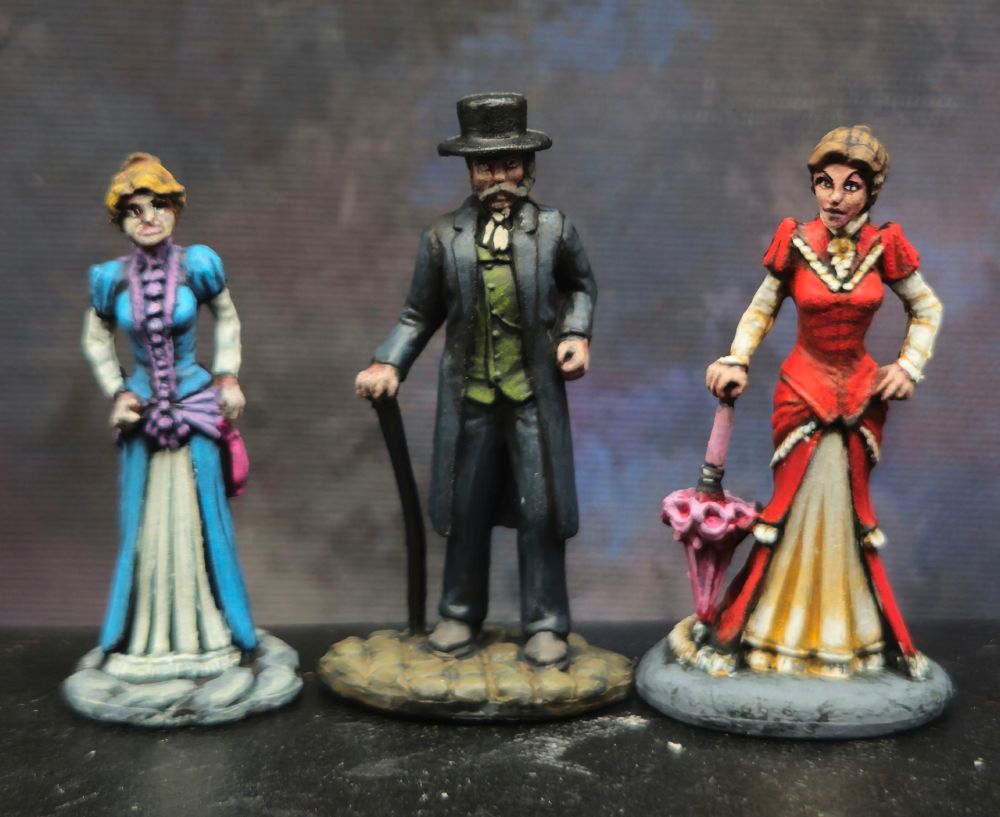 A Victorian gentleman and his 2 lady companions. From left to right. Lady in a blue, white and pink dress with light brown hair. Gentleman in a black suit with a green waistcoat, lady in a red and cream colored dress with a pink parasol. 