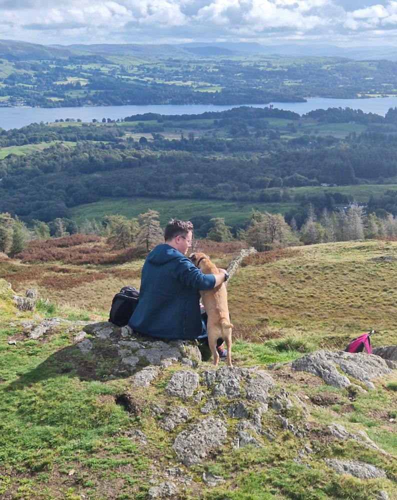 A brown border terrier dog having a cuddle with his male owner on a large hill . A large lake and green hills can be seen in the distance. 