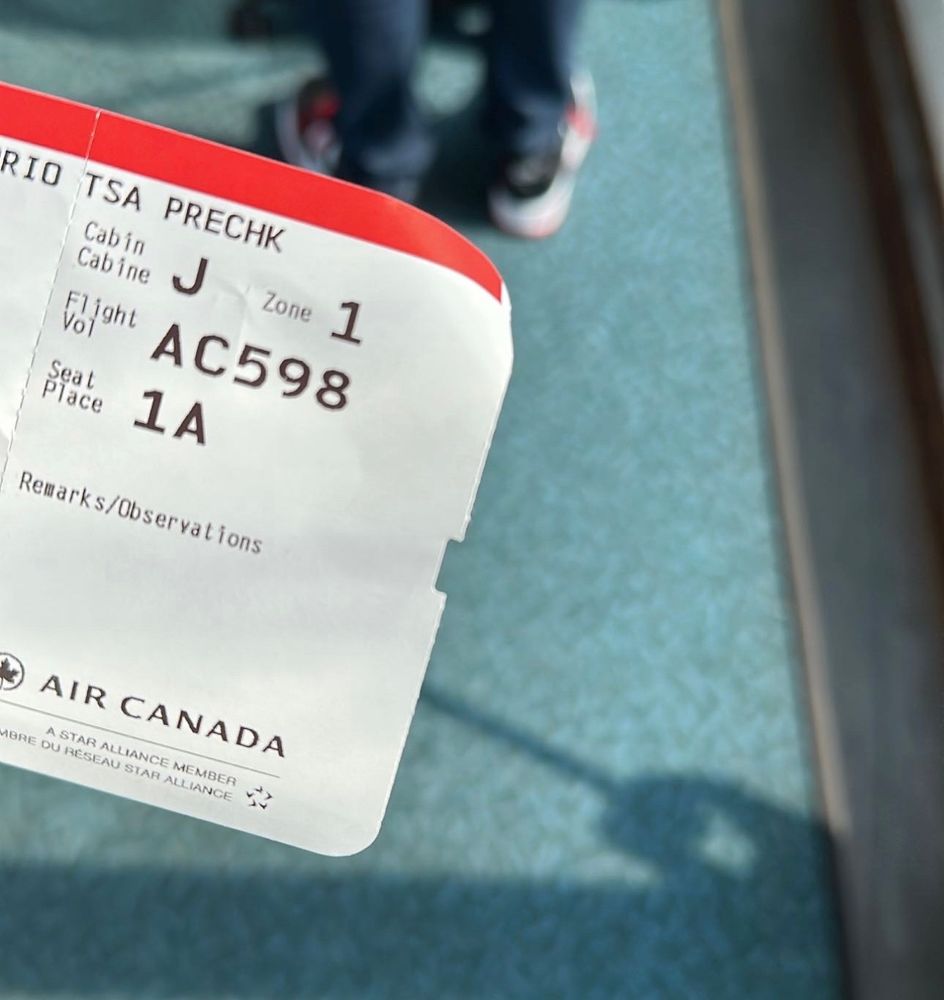 An airplane boarding pass is seen on the left of the photo frame, the boarding pass (acquired with travel points) i dictates TSA PRECHECK, zone 1, seat 1A. Behind the boarding pass is the floor of the walkway to boarding the plane