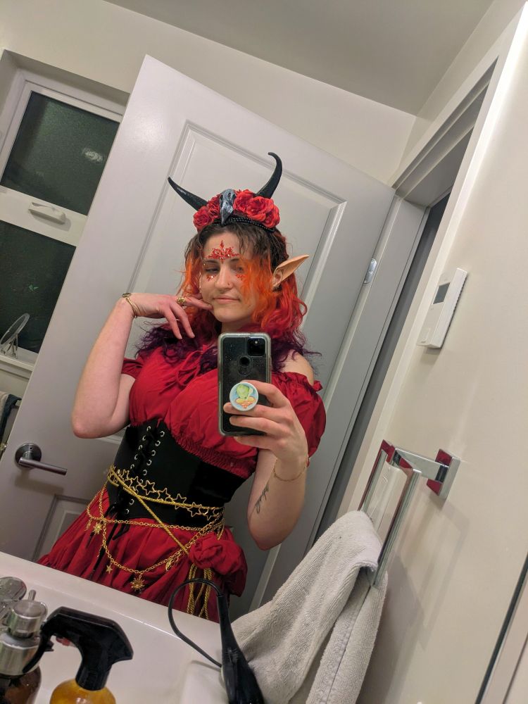 mirror selfie of me, a pale person with hair dyed in an ombre of orange, red, and purple wearing a red dress, elf ears, horns, and a corset draped in gold chain belts