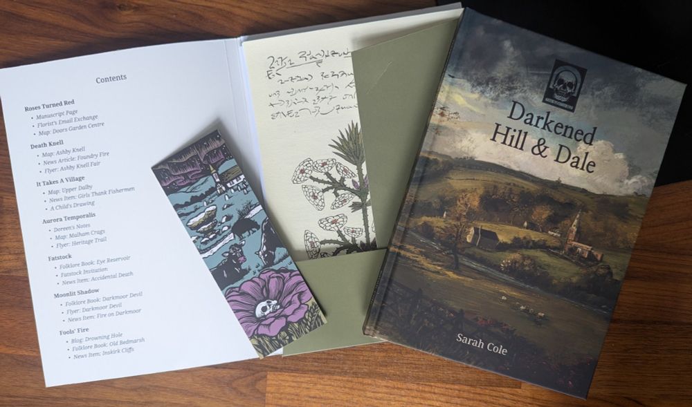 A photo of the book Darkened Hill & Dale by Patchwork Fez Games, sat on a table next to a stylised bookmark and a booklet of handouts and listed contents for the collection of British countryside horror scenarios for tabletop roleplaying games.