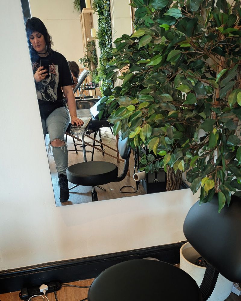 Photo of Lukalock taken of herself sat opposite a mirror, sat on a tattoo bench. She is wearing a black shirt with ripped jeans and black boots.