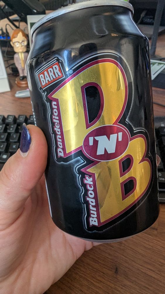 Photo of Lukalock's hand with dark blue painted fingernails, holding a can of "Dandelion & Burdock" soda. In the background is a wooden desk with a keyboard and a bobblehead figure of Dwight from The Office TV show.