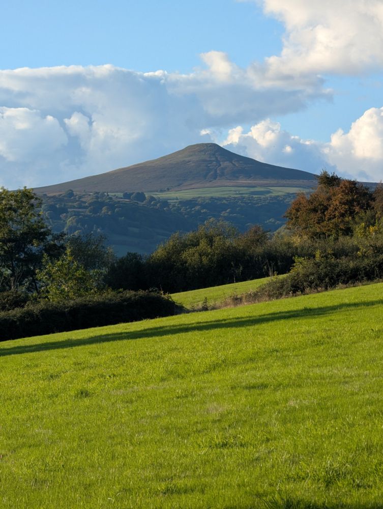 The peak of a hill against the blue sky. Fields and trees in foreground. 