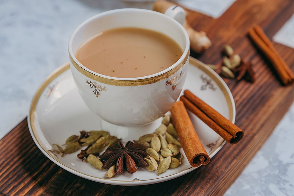 A cute gilded mug of Masala chai with the spice ingredients on the saucer and cutting board its all on - you can see a bit of ginger in the background behind the cup as well as cinnamon sticks, Star Annis, and many other spices. The tea itself is milky with cream.