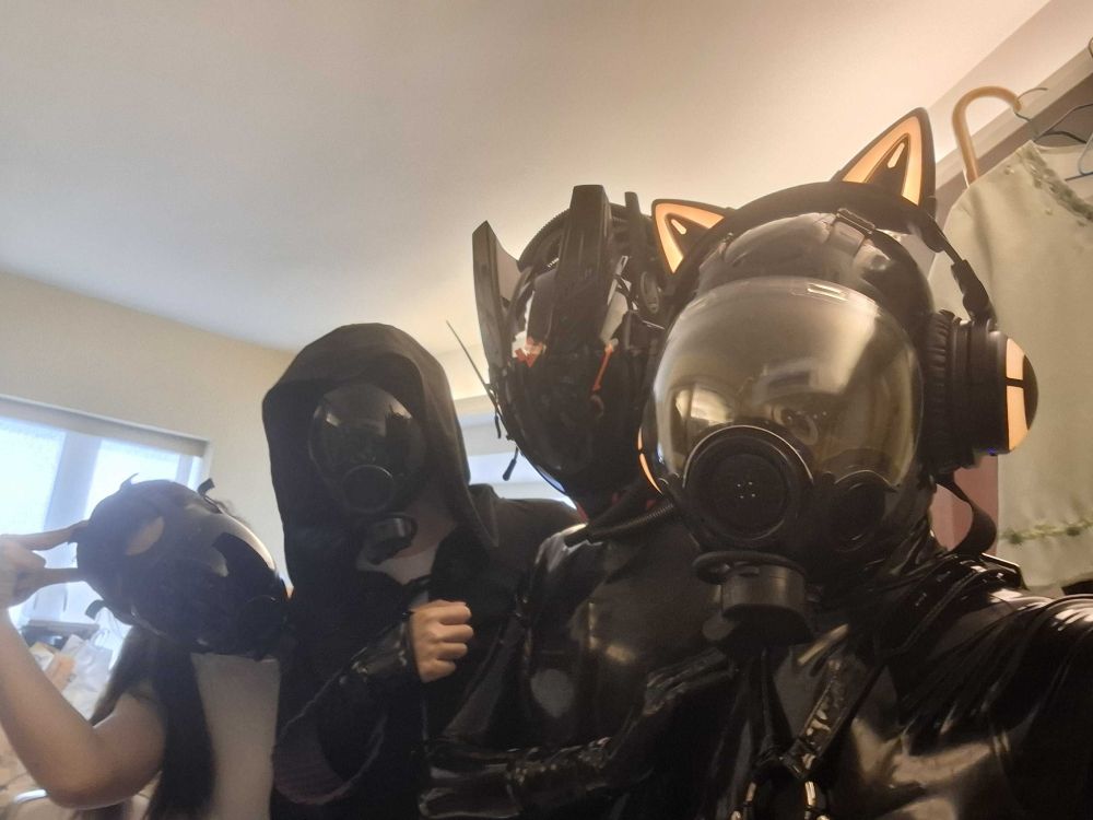 A selfie of four beings in various states of partial dronification, taken inside the HumidHive. From left to right: 5111 is forming a cat ear upon its head with its right arm; it is wearing a tinted gas mask and no other drone gear. 0907 is wearing a tinted gas mask, latex fingerless gloves, and a hoodie covering the rest of its visible body. Ivan has his arms crossed. 4543 is in full HexCorp gear, with its cat ear headphones coloured orange.