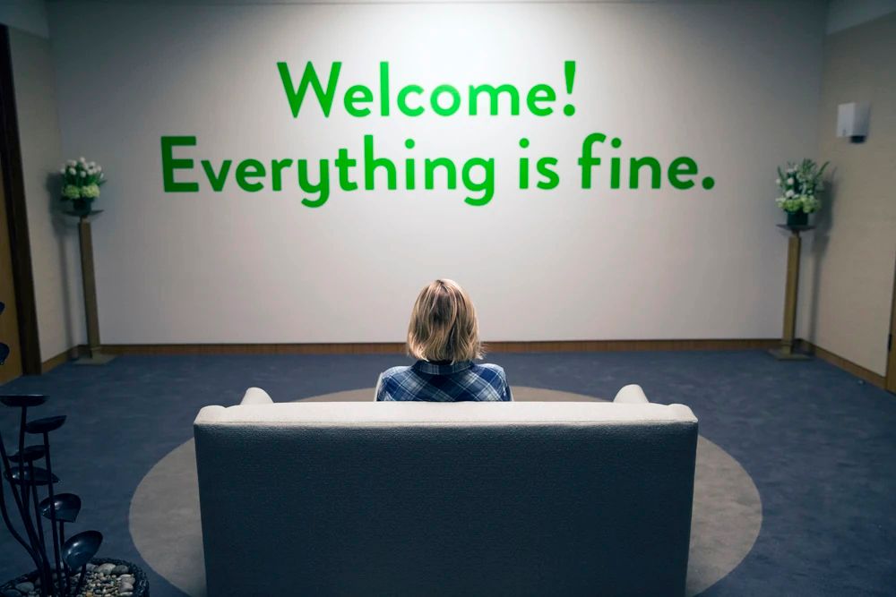 Still from The Good Place. Eleanor sits on a couch in front of a green sign that reads "Welcome! Everything is fine."