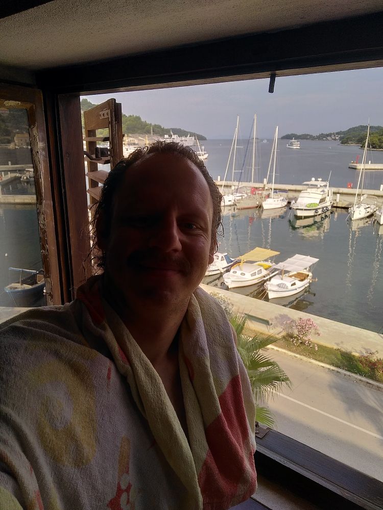 Selfie vor einem offenen Fenster, dahinter ein Hafen mit glattem Wasser und ein paar kleinen Booten.