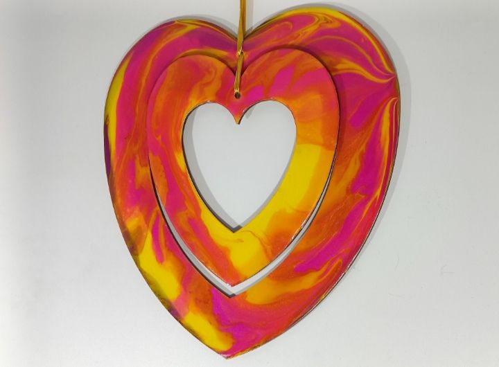 Two hearts hung together on a golden yellow ribbon against a white background. The hearts are painted in bright pink, yellow and orange abstract flowing patterns