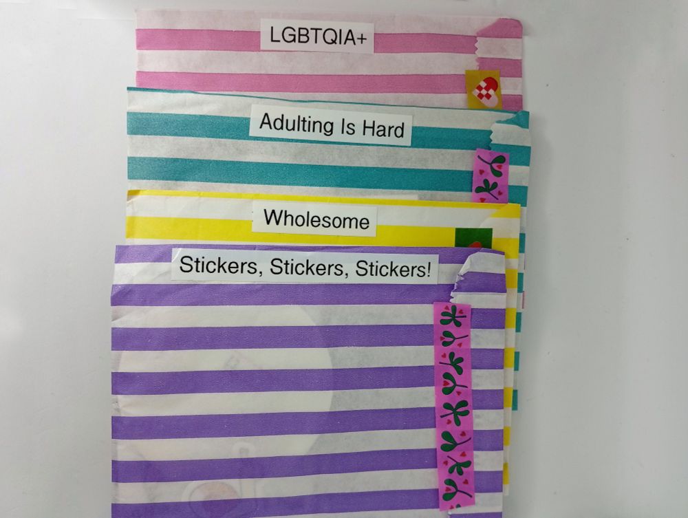 Four colourful striped bags on a white background, sealed with colourful washi tape and labelled with white labels with black font; LGBTQIA+, Adulting Is Hard, Wholesome, and Stickers, Stickers, Stickers!