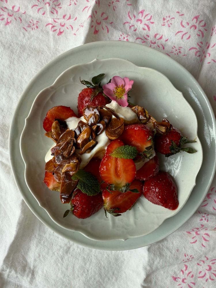 Strawberries, labne and almond brittle on a celadon dish. A strawberry flower and a scattering of mint leaves on top. A white cloth with pink flowers below. 