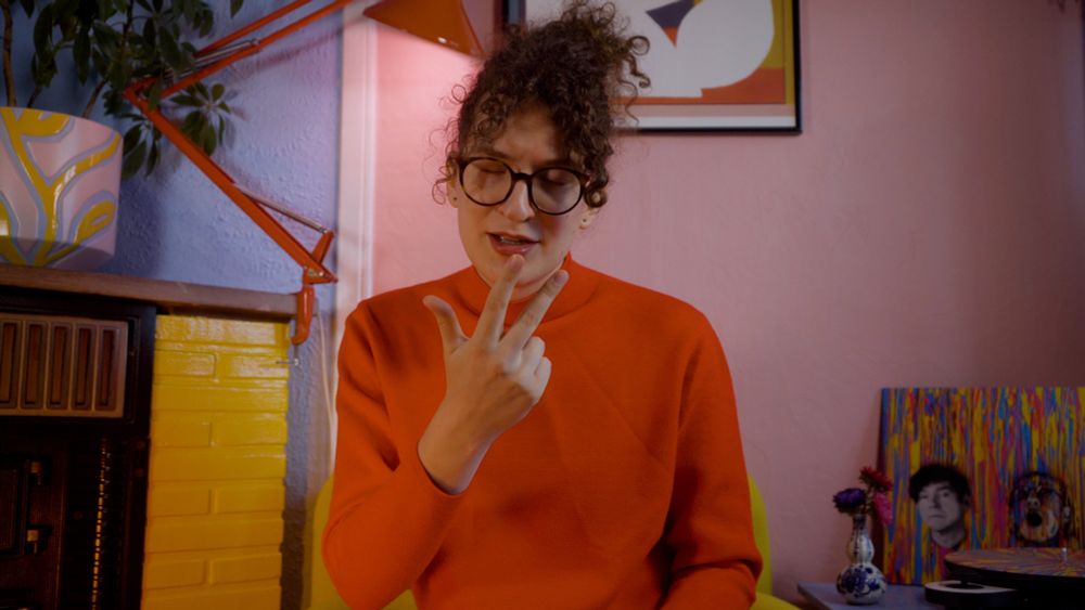 Robin making an amused face at the camera, gesturing three fingers up and clearly in thought. She’s wearing a red sweater sitting in a colourful room.