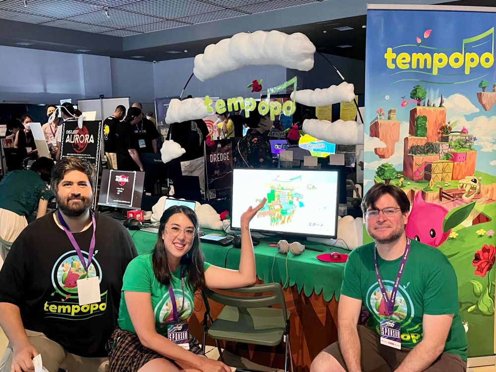 Picture from BitSummit 2023 in front of the Tempopo booth. Three Developers of Tempopo are showing off the hand crafted booth behind. The text 