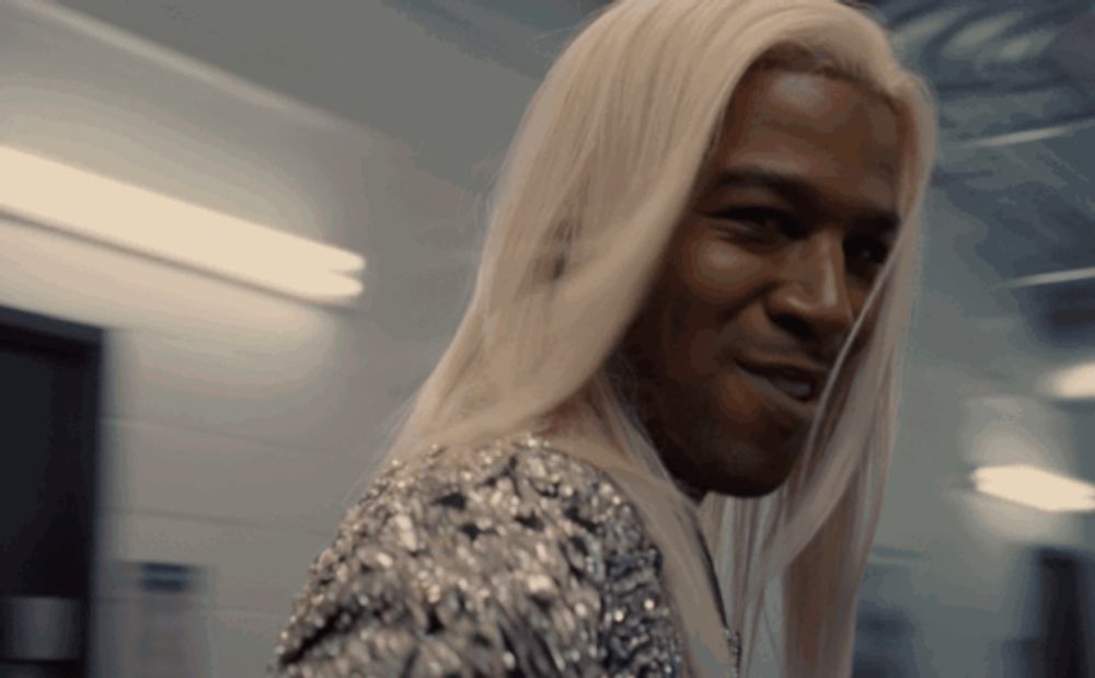 a man with long blonde hair is smiling and wearing a silver dress