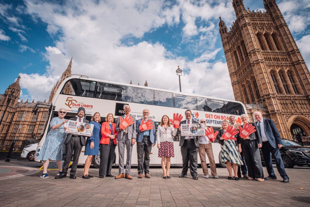 A white bus wrapped in the logos of Catch the Bus Month, Bus Users UK, Dawsongroup and the All-Party Parliamentary Group stands in the middle of Old Palace Yard, Westminster, with the Houses of Parliament behind it, and in front of it stands Claire Walters, Chief Executive of Bus Users UK, Warinder Juss MP, Fleur Anderson MP, Ruth Cadbury MP, John Milne MP, Brian Matthew MP, Alex Mayer MP, Mike Wood MP, Jennie Martin MBE, Chair of Bus Users UK, Lydia Horbury, Director for England, Bus Users UK, Paul Sainthouse, Managing Director of Dawsongroup and International President of CILT, Sir Roger Gale