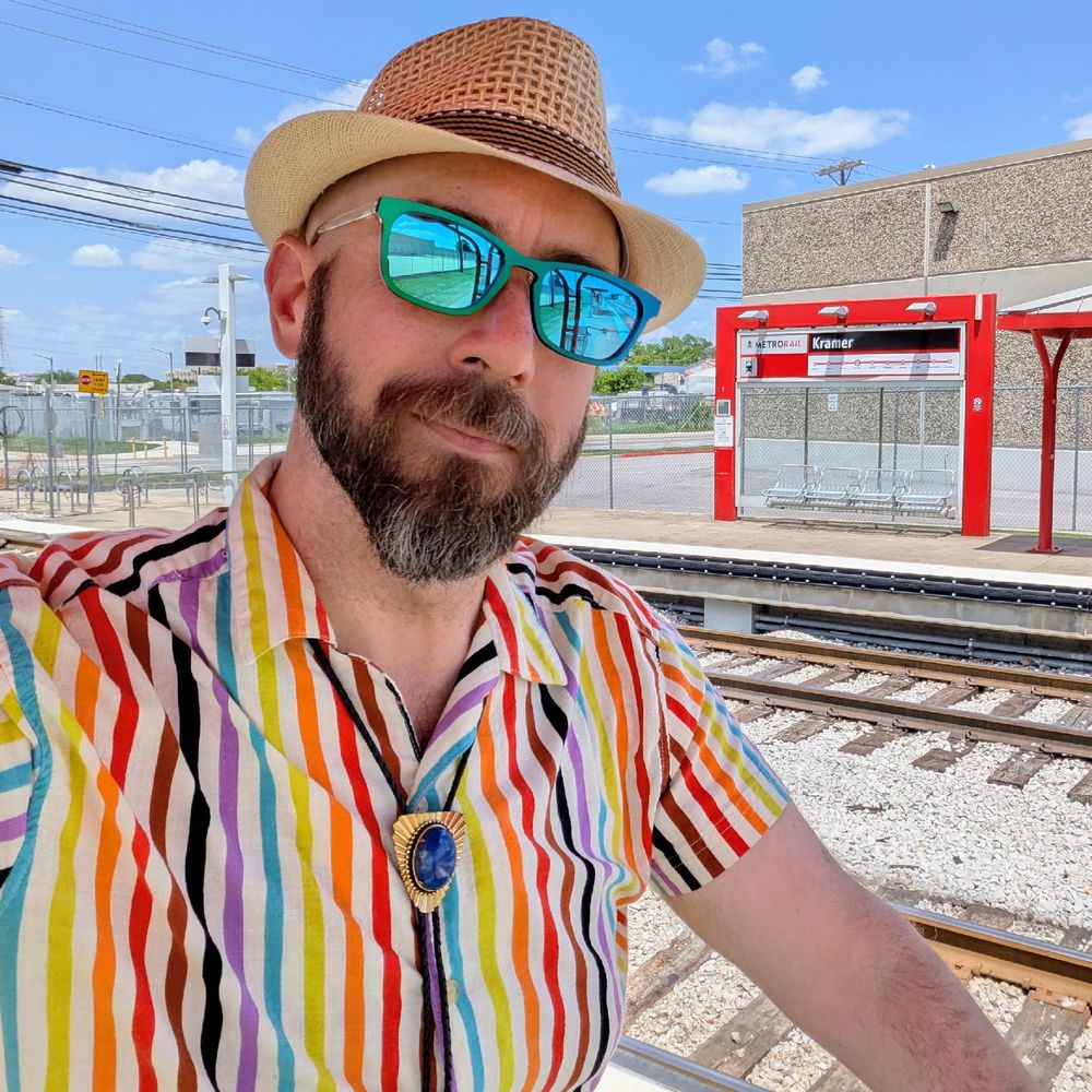 A photo of me, a 47 year old white man with a shaved head, a short cropped brown & grey beard, 

photographed from the chest up

In front of the far side of Kramer Station and it's tracks

wearing

rectangular green-blue metallic acetate sunglasses, a straw fedora, lapis lazuli bolo tie, and a vertical pride rainbow stripesd shirt sleeve button down shirt