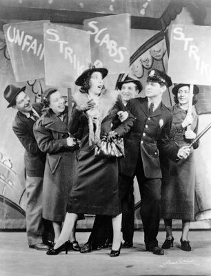 still image from Pins and Needles showing a group of workers on strike with signs saying "strike" "scabs" "unfair" and an actress dressed as a rich lady and an actor dressed as police officer next to them. The still is taken from the number "it's not cricket to picket"