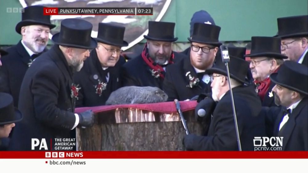 9 men dressed in smart black coats, scarves, and top hats, gather around a tree stump, a groundhog it atop. A white bearded man at the front holds a staff and talks to the groundhog apparently in groundhogese. 