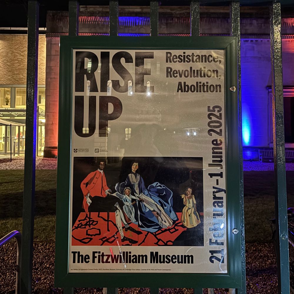 A poster fixed to the railings in front of a museum, lit up in blue and yellow light. The poster reads: Rise Up Resistance, Revolution, Abolition. 