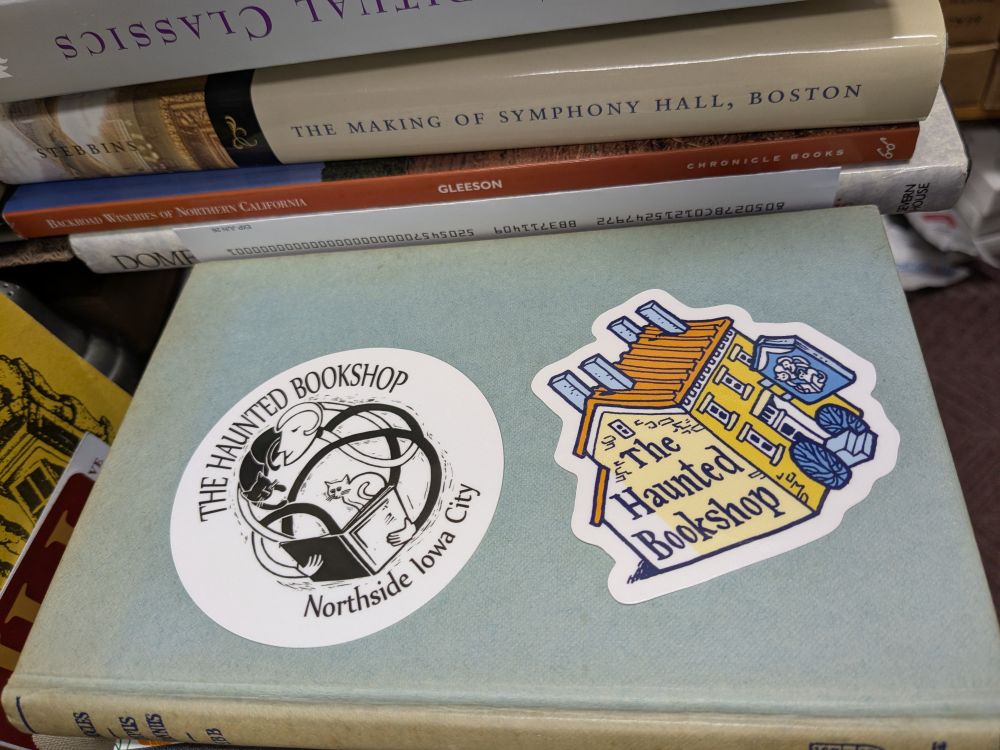 Two three inch stickers, one with this bookshop's name and location (The Haunted Bookshop / Northside Iowa City) around the black and white logo depicting a reader with a book and two nosy cats. The other sticker is a drawing of the shop as seen from Gilbert Street, with our yellow stone walls, red roof, and Greek Revival era windows. The drawing is by Kevin Cannon. The rest of the picture is, of course, stacks of secondhand books.