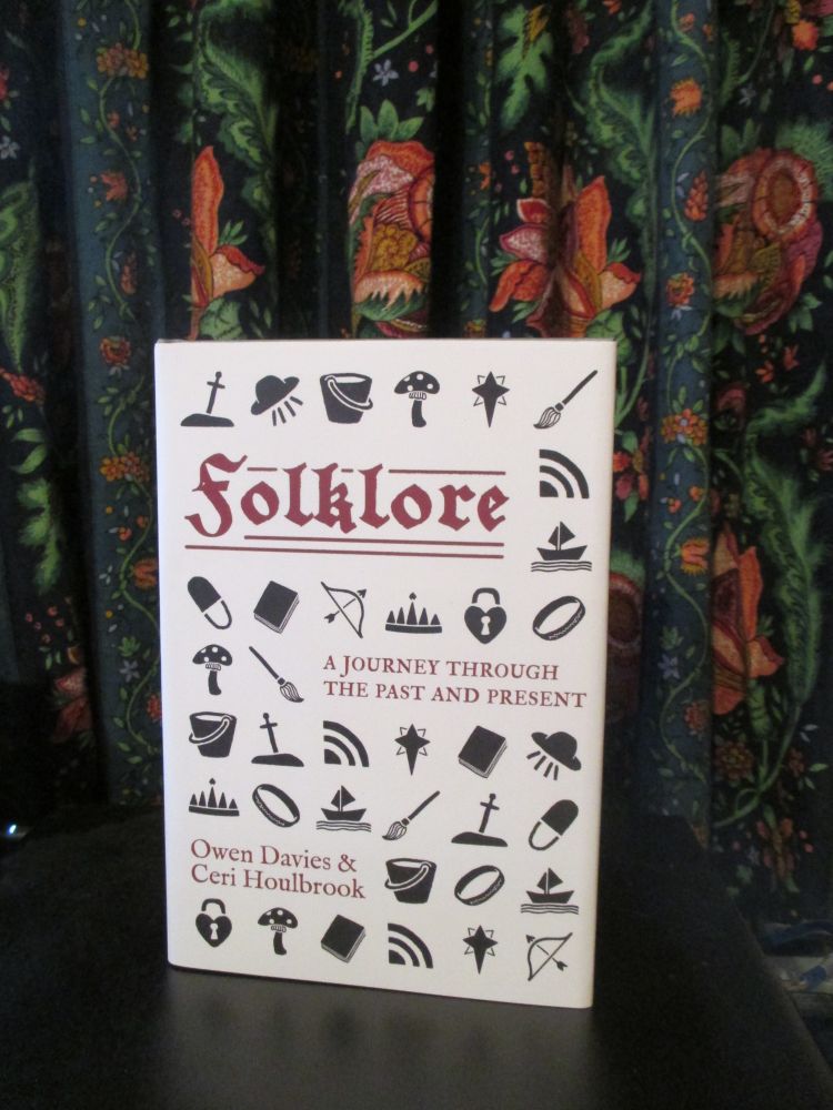 Light grey cover of Folklore: A Journey Through the Past and Present by Owen Davies and Ceri Houlbrook, showing folklore symbols like a lock, bow and arrow, mushroom, broom, and ring. The book stands in front of a brightly colored curtain.